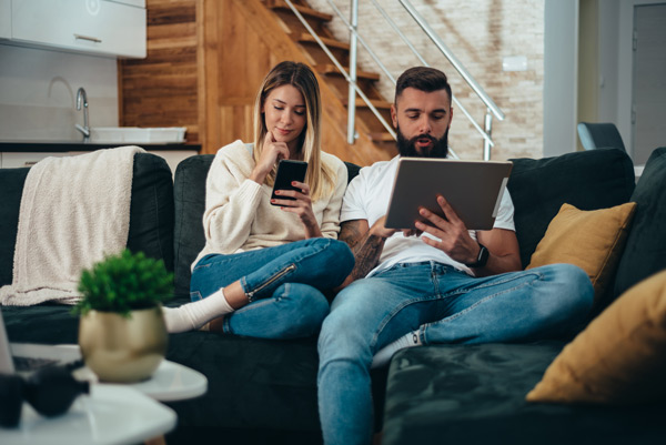 casal sentado em sofá, mulher utilizando smartphone e homem utilizando tablet