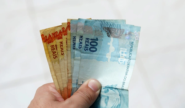 Mão segurando notas de 100 e 20 reais.
